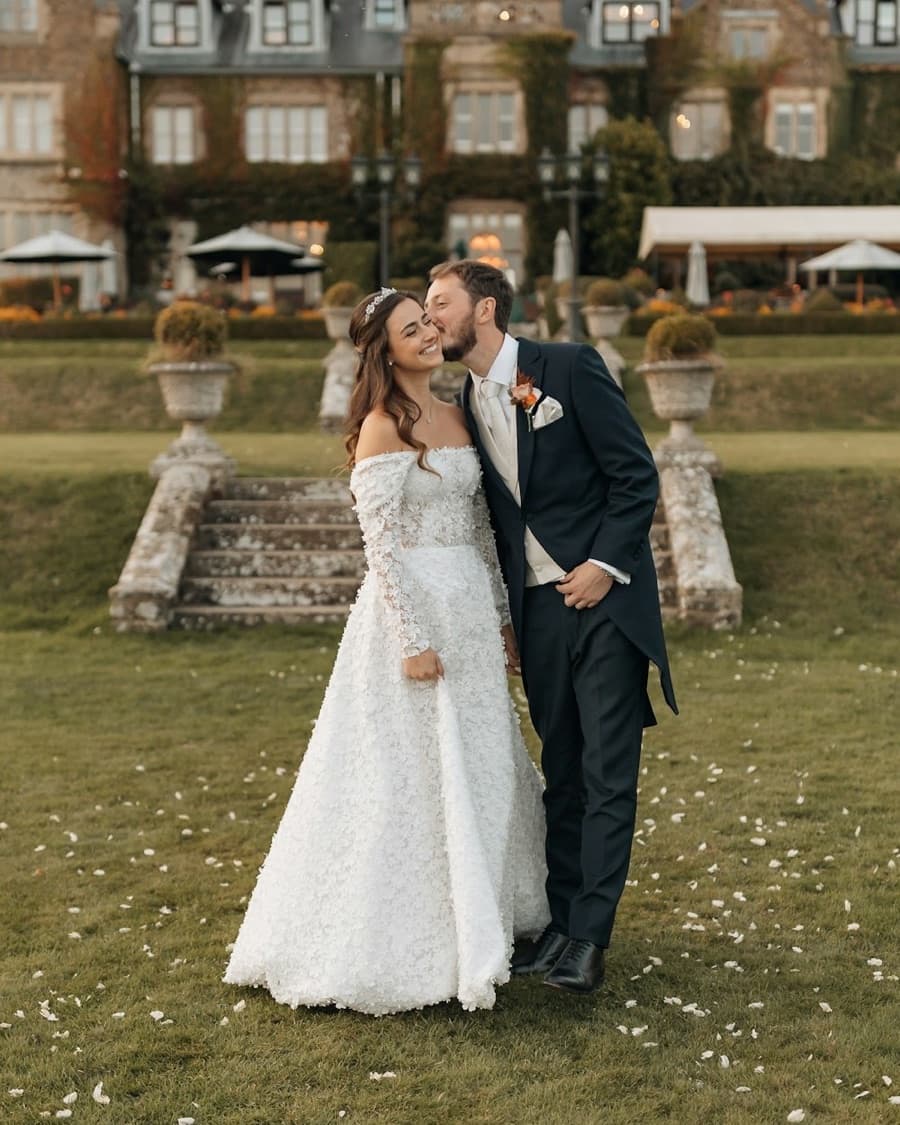 Mina in an off-the-shoulder lace gown, laughing as Curtis kisses her cheek on the lawn below a country house
